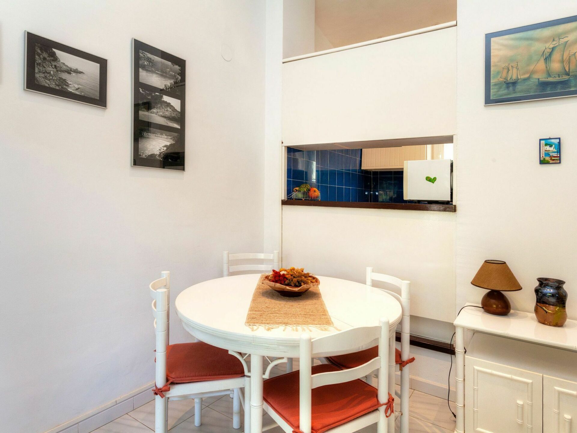 A dining area with a round white table, white chairs, and a small side table with a lamp and decorative items.