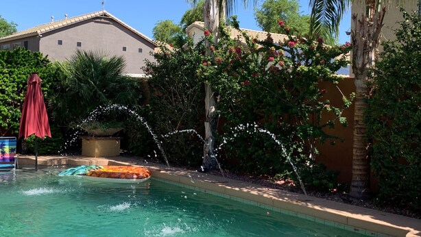 A clear swimming pool with a water fountain, surrounded by palm trees and a building with a tiled roof. - VISIT ARIZONA IN STYLE                   ENJOY A HEATED POOL (Goodyear)