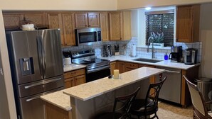 A modern kitchen with stainless steel appliances, wooden cabinets, and a large island with bar stools. - VISIT ARIZONA IN STYLE                   ENJOY A HEATED POOL (Goodyear)