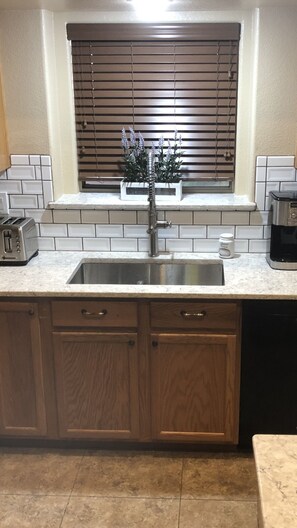 A kitchen with a stainless steel sink, wooden cabinets, a window with blinds, and a toaster. - VISIT ARIZONA IN STYLE                   ENJOY A HEATED POOL (Goodyear)