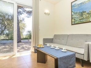 Una habitación con sofá, una mesa con tazas y pasteles, un cuadro en la pared y vistas a través de un ventanal.