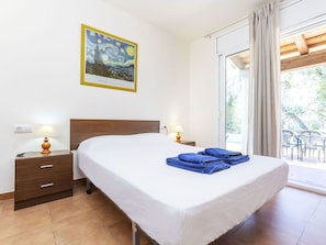 Una habitación de hotel con una cama, dos mesitas de noche con lámparas, una mesita auxiliar, una ventana con cortinas y un cuadro en la pared.