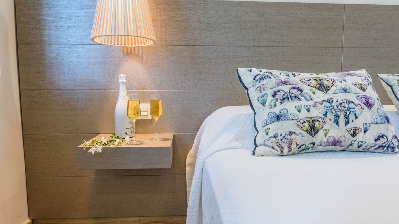 Una cama bien hecha con un cojín floral, una mesita de noche con una botella y vasos, y una lámpara de pared.