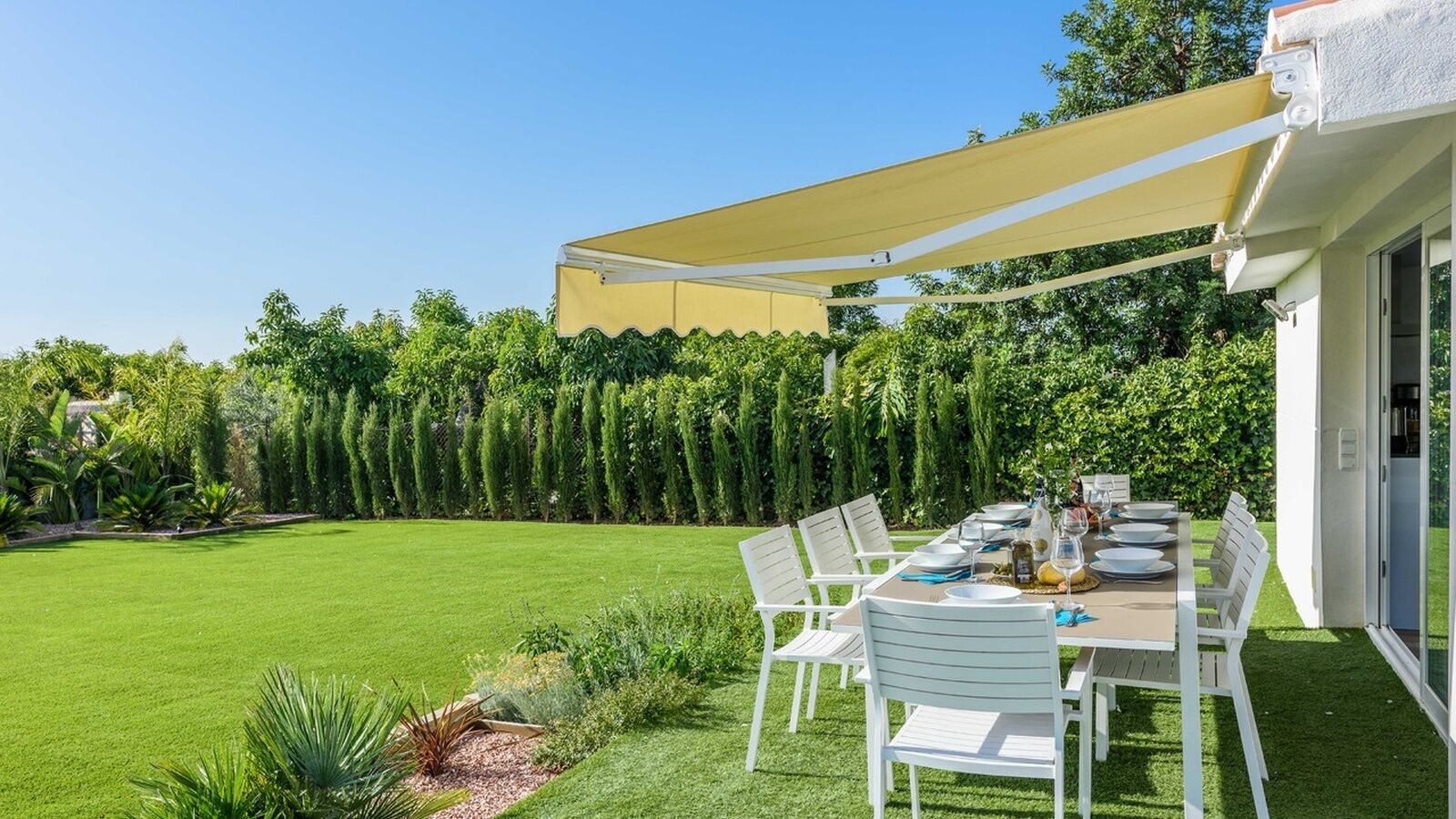 Un área de comedor al aire libre bien organizada, con una mesa lista para cuatro personas, rodeada de sillas blancas, bajo un toldo amarillo.