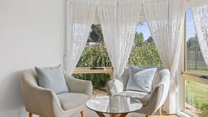 A cozy seating area with two armchairs and a glass table, surrounded by sheer white curtains and a view of greenery outside.