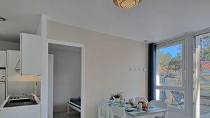 A compact kitchen with white cabinets and a built-in stove, a dining area with a table and chairs, and a large window offering a view of the outdoors. - Lovely apartment in villa for 4 guests with WIFI, pool, TV, terrace and pets allowed (Saint-hilaire-de-riez)