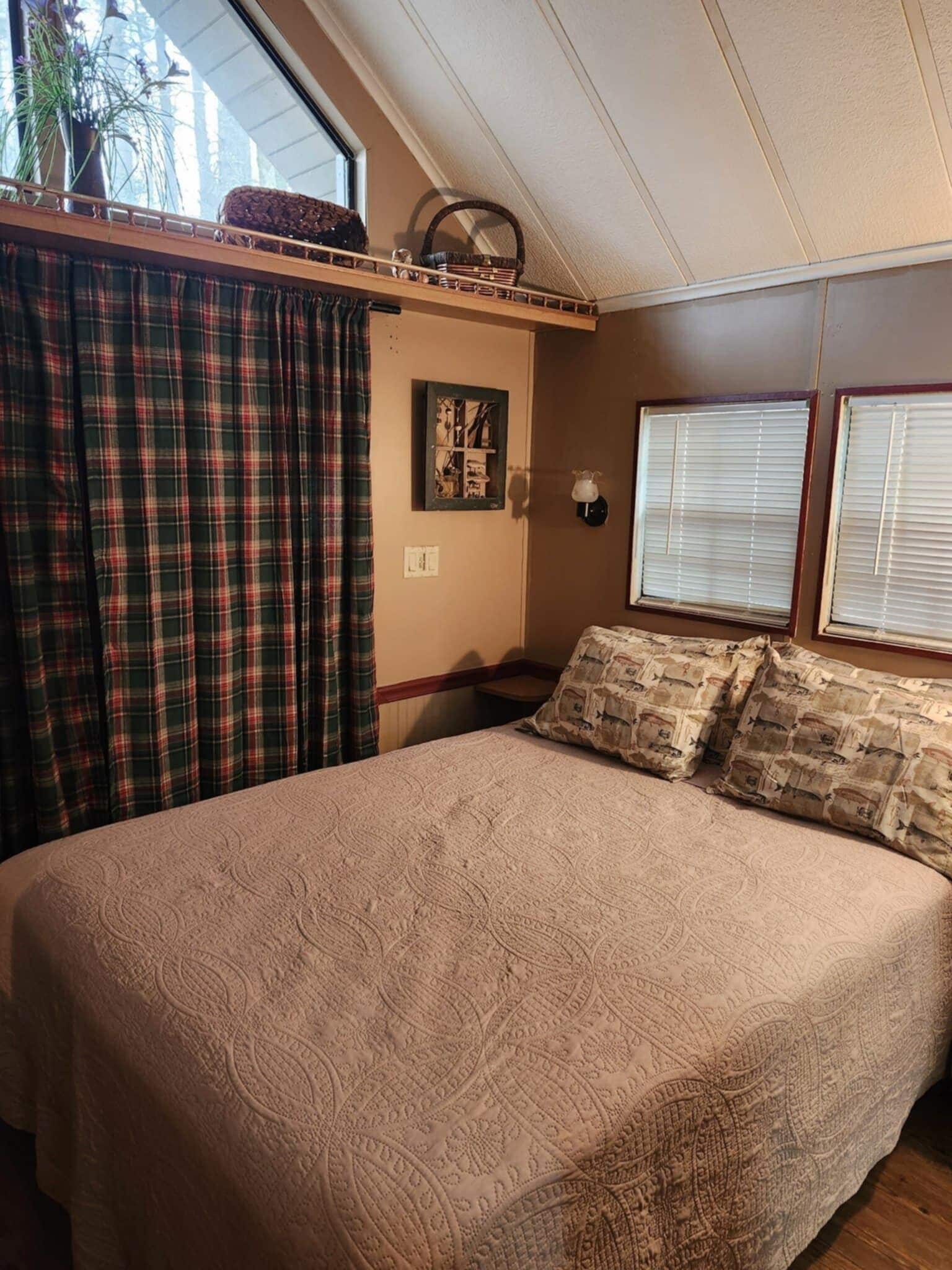 A bedroom with a large bed, plaid curtains, a window with blinds, and a small table with a lamp.