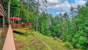 A wooden deck with a red umbrella, overlooking a forested area with a stream.