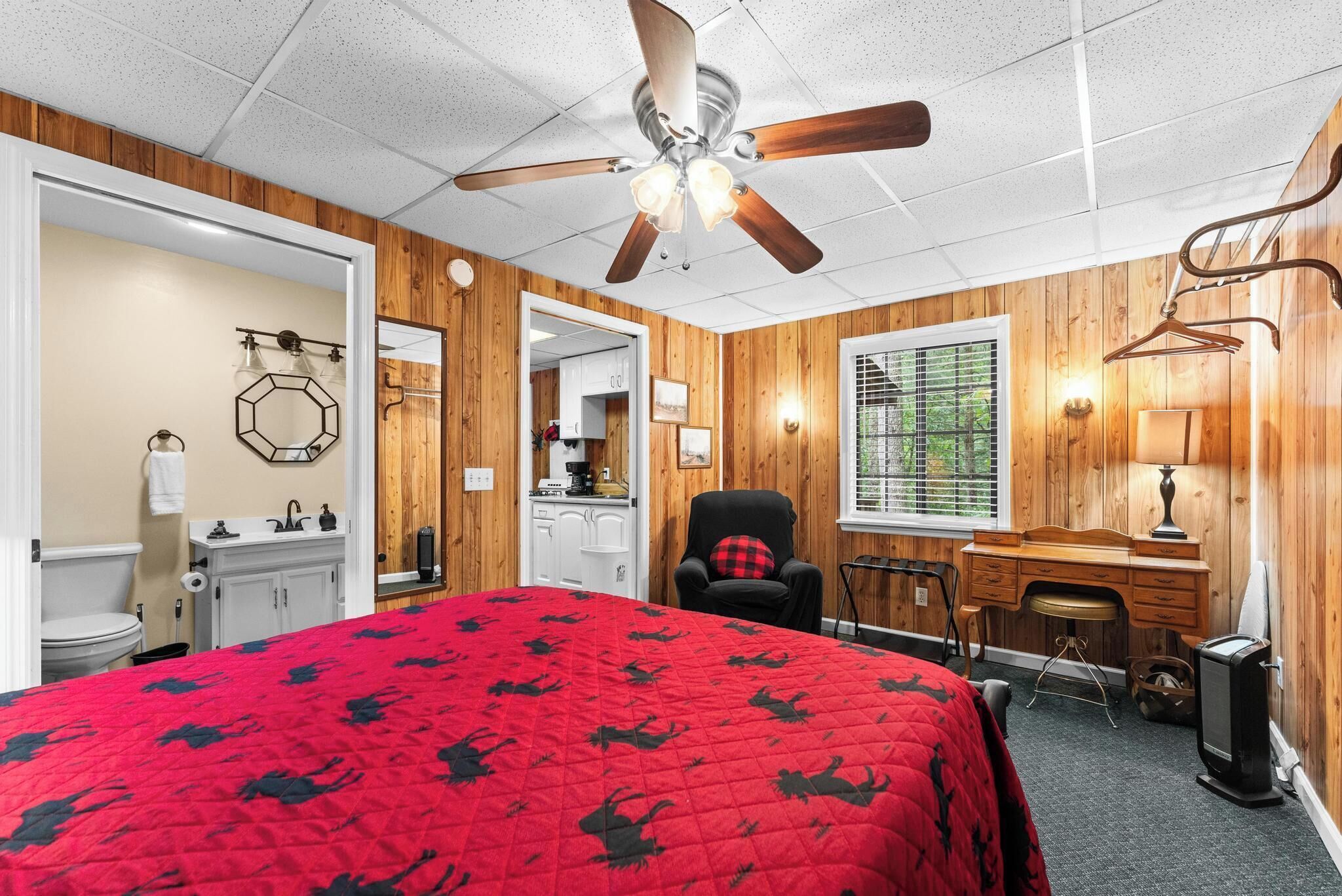 A bedroom with a red bedspread, wooden walls, a ceiling fan, a desk, and a television.