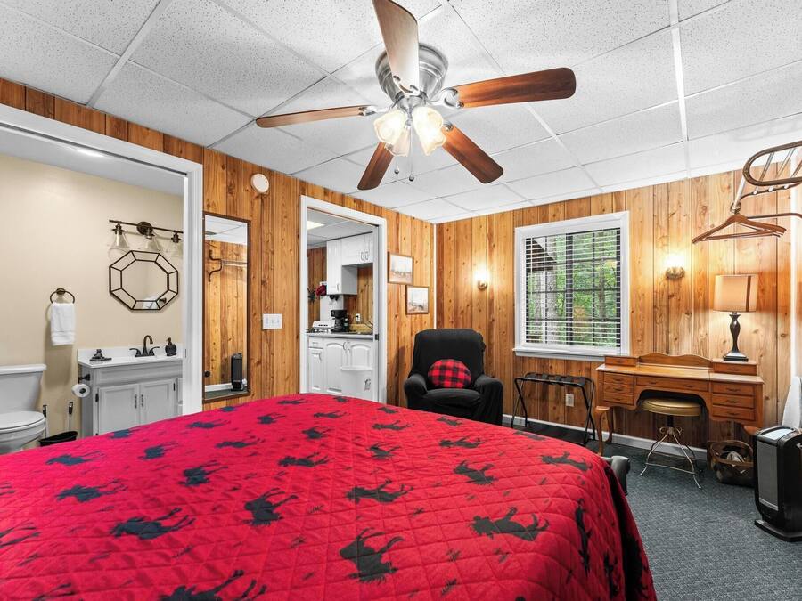 A bedroom with a red bedspread, wooden walls, a ceiling fan, a desk, and a television.