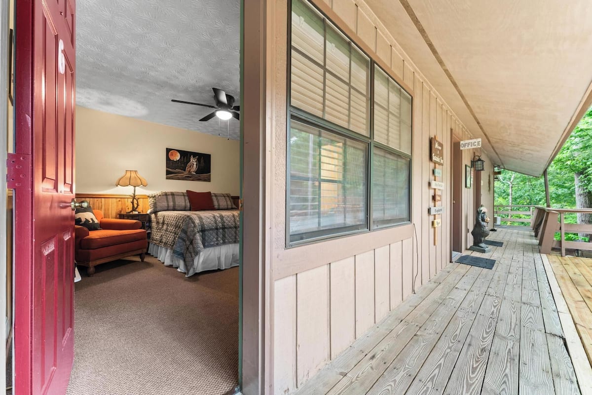 A wooden exterior with a covered porch, a red door, and a bed with a lamp in the adjacent room.