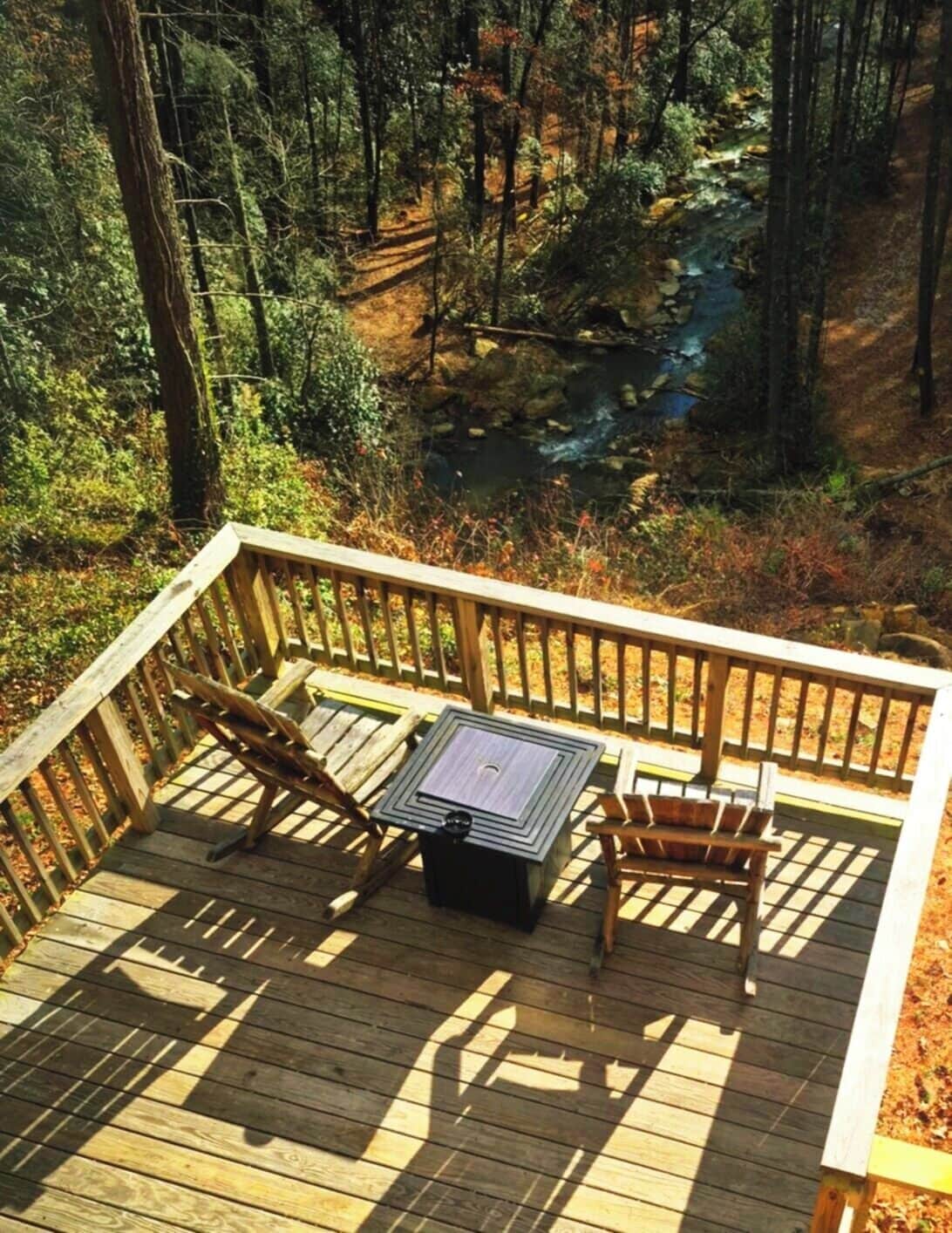 A wooden deck with a table and chairs overlooking a forested area and a stream.