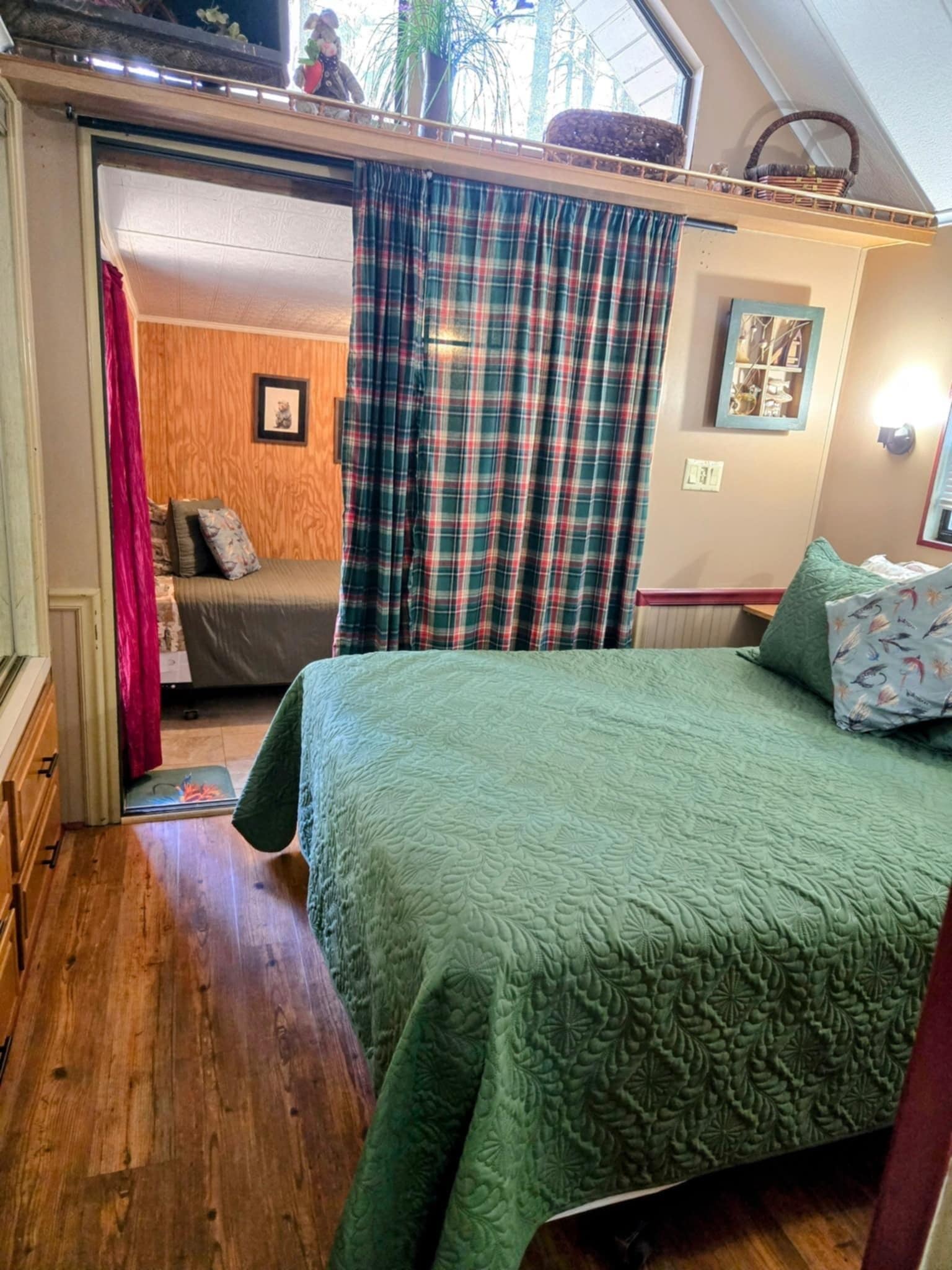 A cozy bedroom with a green quilted bedspread, a wooden floor, a window with plaid curtains, and a small table with a teddy bear.