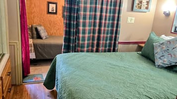 Un dormitorio acogedor con una colcha verde acolchada, suelo de madera, una ventana con cortinas escocesas y una mesita de noche con un osito de peluche.