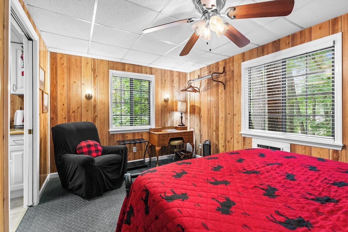 A bedroom with wooden walls, a ceiling fan, a bed with a red bedspread, a desk, a chair, and a window with blinds.