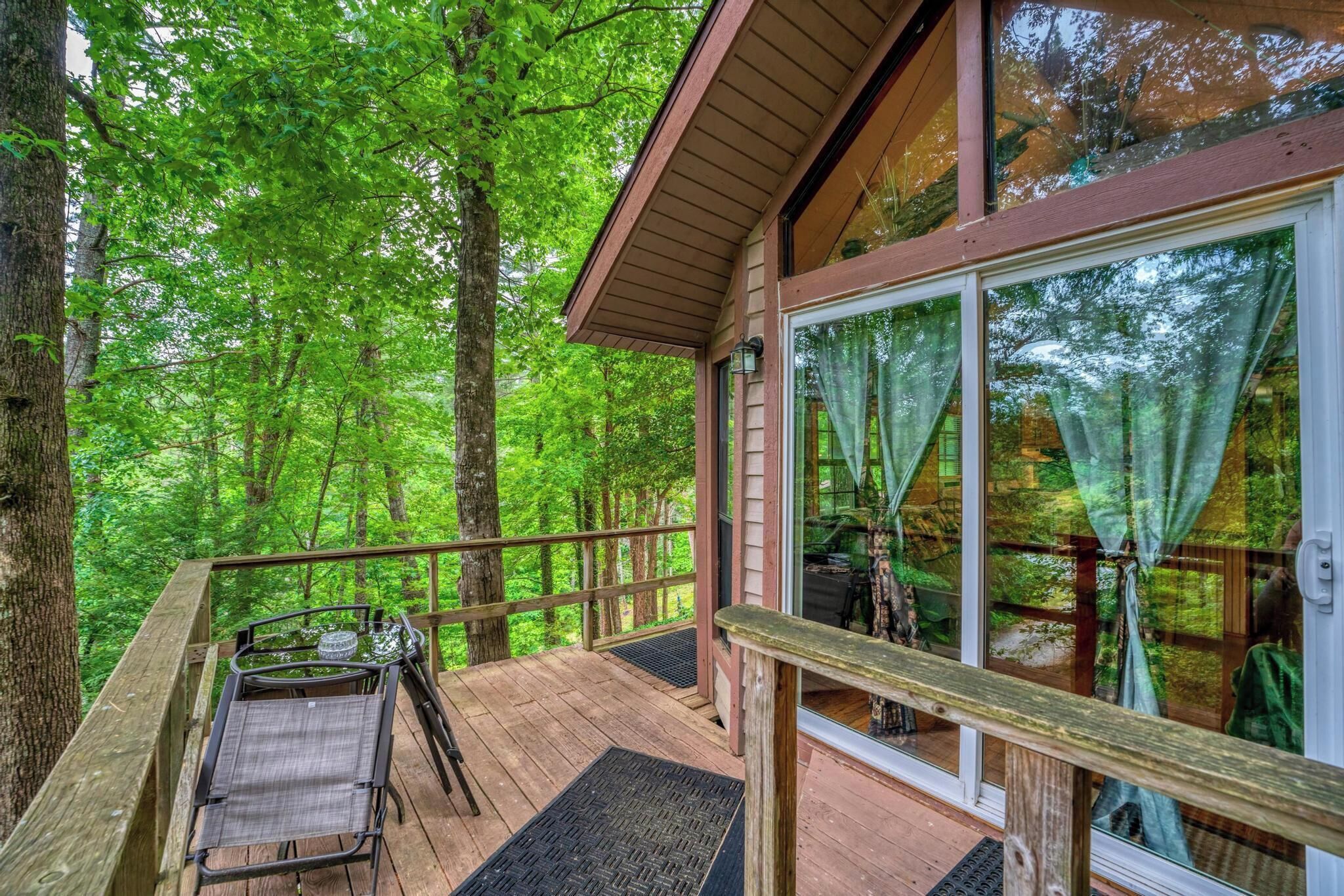Una terraza de madera con un ventanal de vidrio que da a una habitación, rodeada de un bosque.