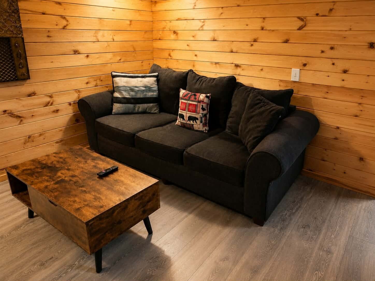 A cozy room with a wooden wall, a dark-colored sofa with multiple cushions, and a small wooden table with a remote control on it.