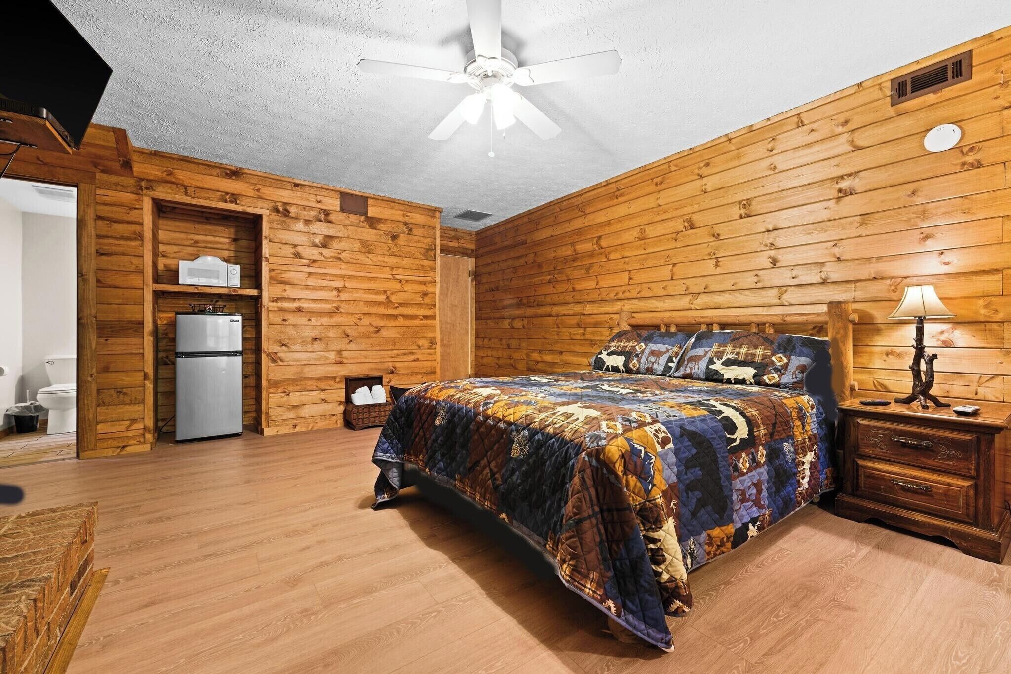 A bedroom with a wooden floor, a bed with a quilt, a ceiling fan, a nightstand, and a small table with a lamp.