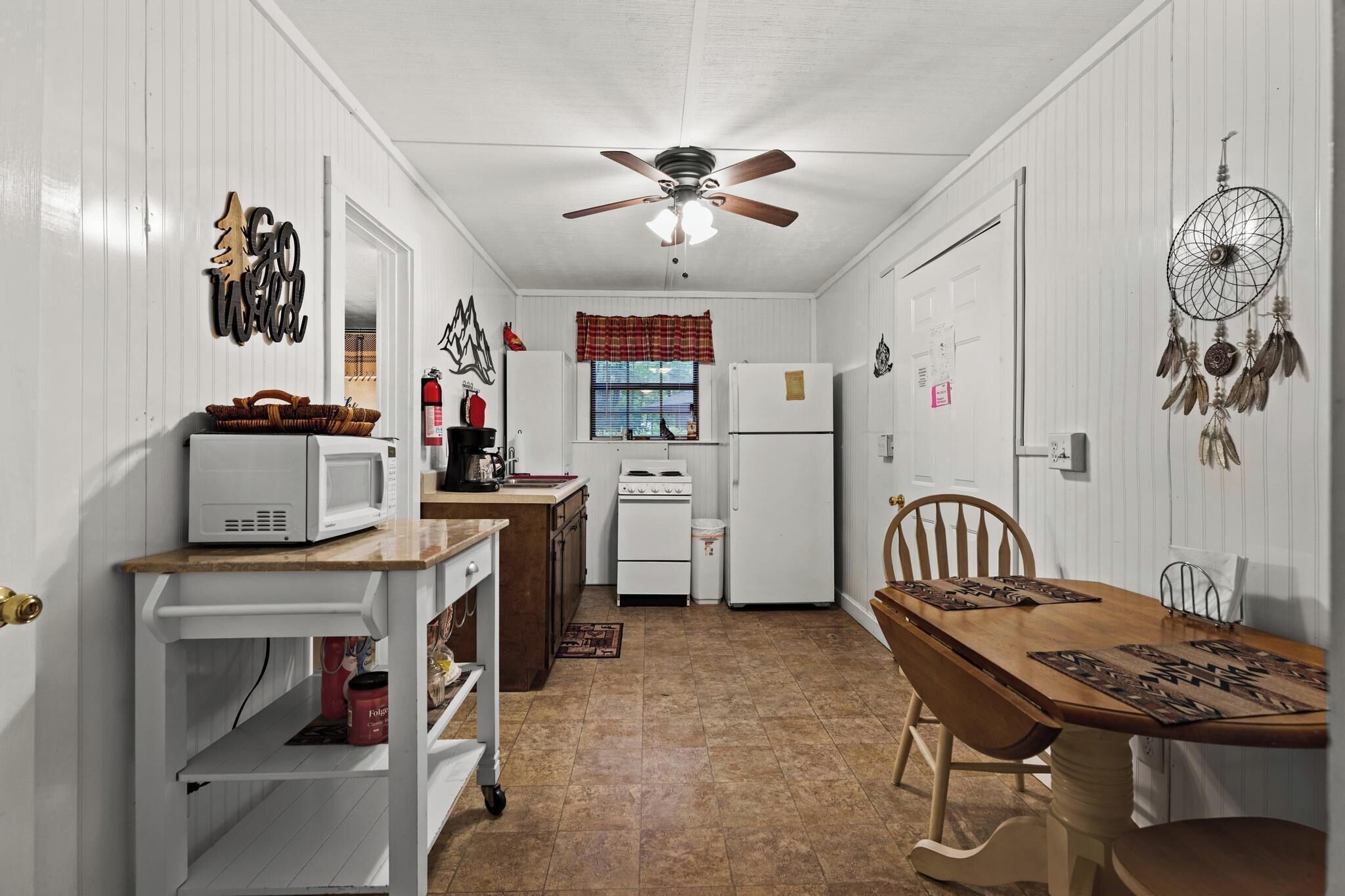 A compact kitchen with white cabinets, a microwave, a stove, and a refrigerator. There is a dining table with chairs and a ceiling fan.
