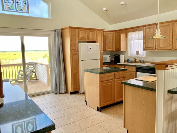 A kitchen with wooden cabinets, a refrigerator, a microwave, and a dishwasher. There is a sliding glass door leading to a balcony with a view of a field.