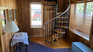 Interior de una cabaña de madera con una escalera de caracol, una mesa con mantel de encaje y un banco.