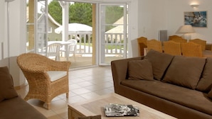 Ein Wohnzimmer mit braunem Sofa, Korbstuhl und Couchtisch mit einer Zeitschrift. Eine Glastür führt zu einem Außenbereich mit einem weißen Sonnenschirm und Stühlen.