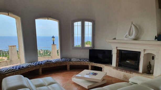 Un salon avec un grand canapé, un téléviseur et une cheminée. De grandes fenêtres offrent une vue sur la mer.