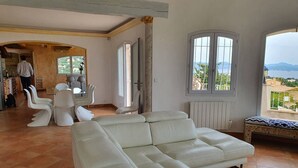 Un vaste salon doté d’un canapé en cuir blanc, d’un coin repas avec des chaises blanches et de grandes fenêtres offrant une vue sur la mer.