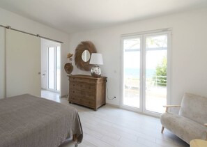 Een slaapkamer met een bed, een dressoir, een stoel en een groot raam met uitzicht op zee.