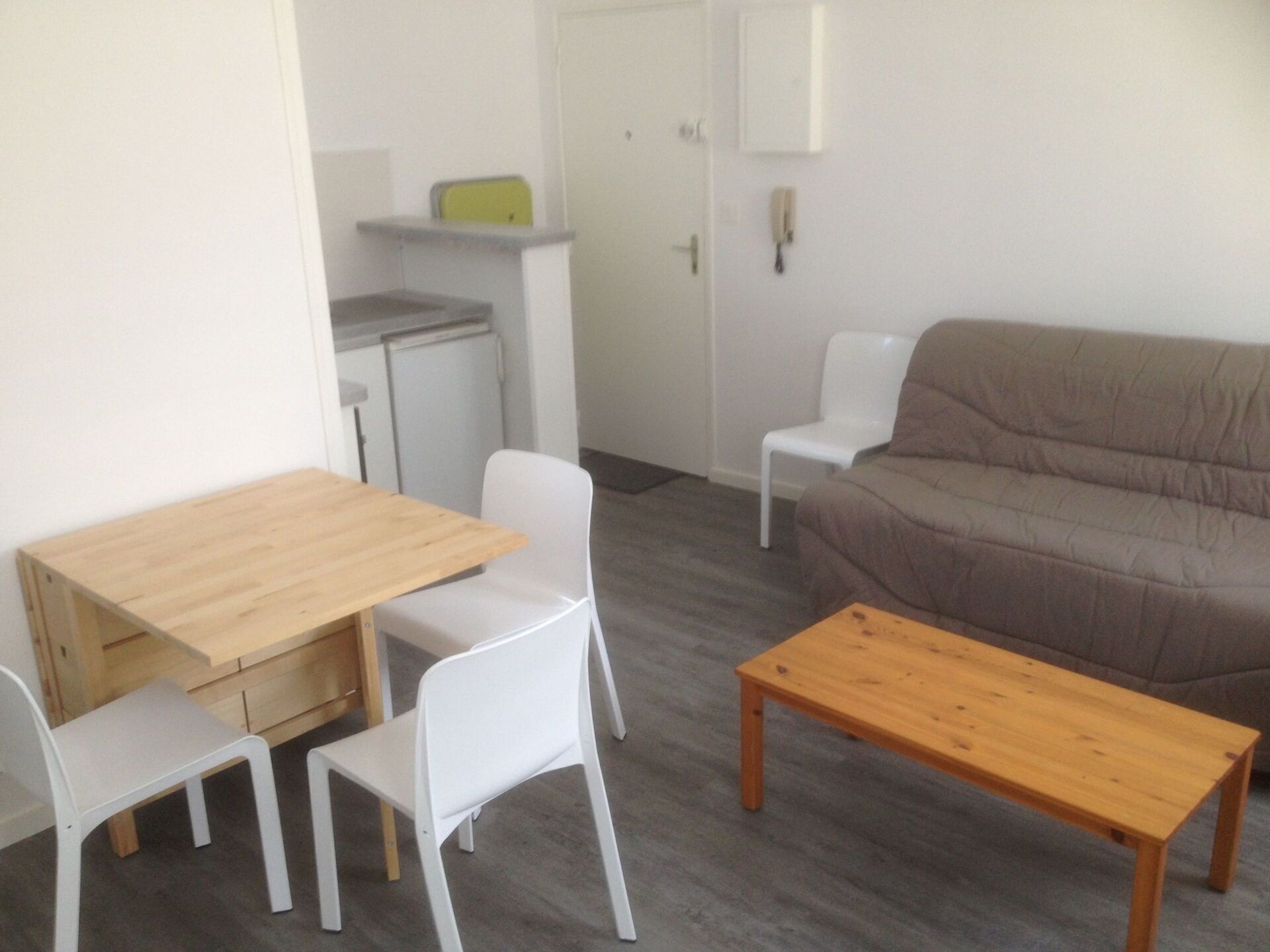 A small, modern living room with a wooden table, two white chairs, a brown sofa, and a small kitchenette in the background.