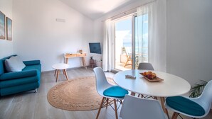 Een moderne woonkamer met een blauwgroene bank, een ronde witte tafel met stoelen, een groot raam met gordijnen en uitzicht op de omgeving.