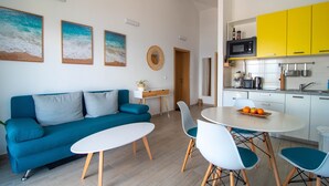 Een moderne woonkamer met een blauwe bank, een witte salontafel en teal stoelen. In de achtergrond is een keuken zichtbaar met gele kastjes en een wit aanrecht.