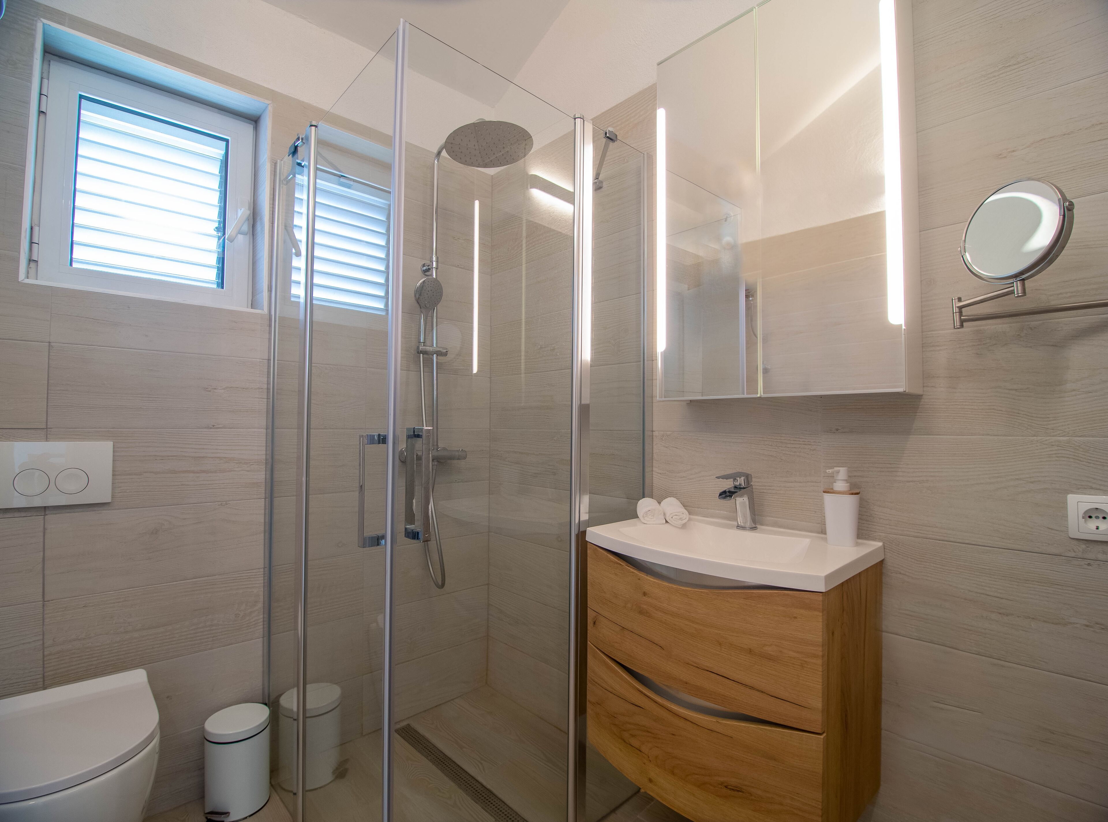 Ein modernes Badezimmer mit einer gläsernen Duschkabine, einem Holzkosmetikschrank, einer Toilette und einem Fenster mit Jalousien.