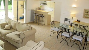 A modern living room with a glass dining table and chairs, a beige sofa, and a kitchen in the background. - Beautiful apartment in villa for 8 guests with private pool, TV, terrace and pets allowed (Pont-de-larn)