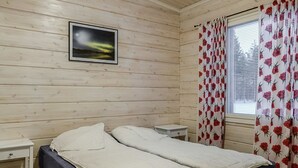 A room with two beds, a small bedside table, a nightstand, a window with floral curtains, and a framed picture on the wall.