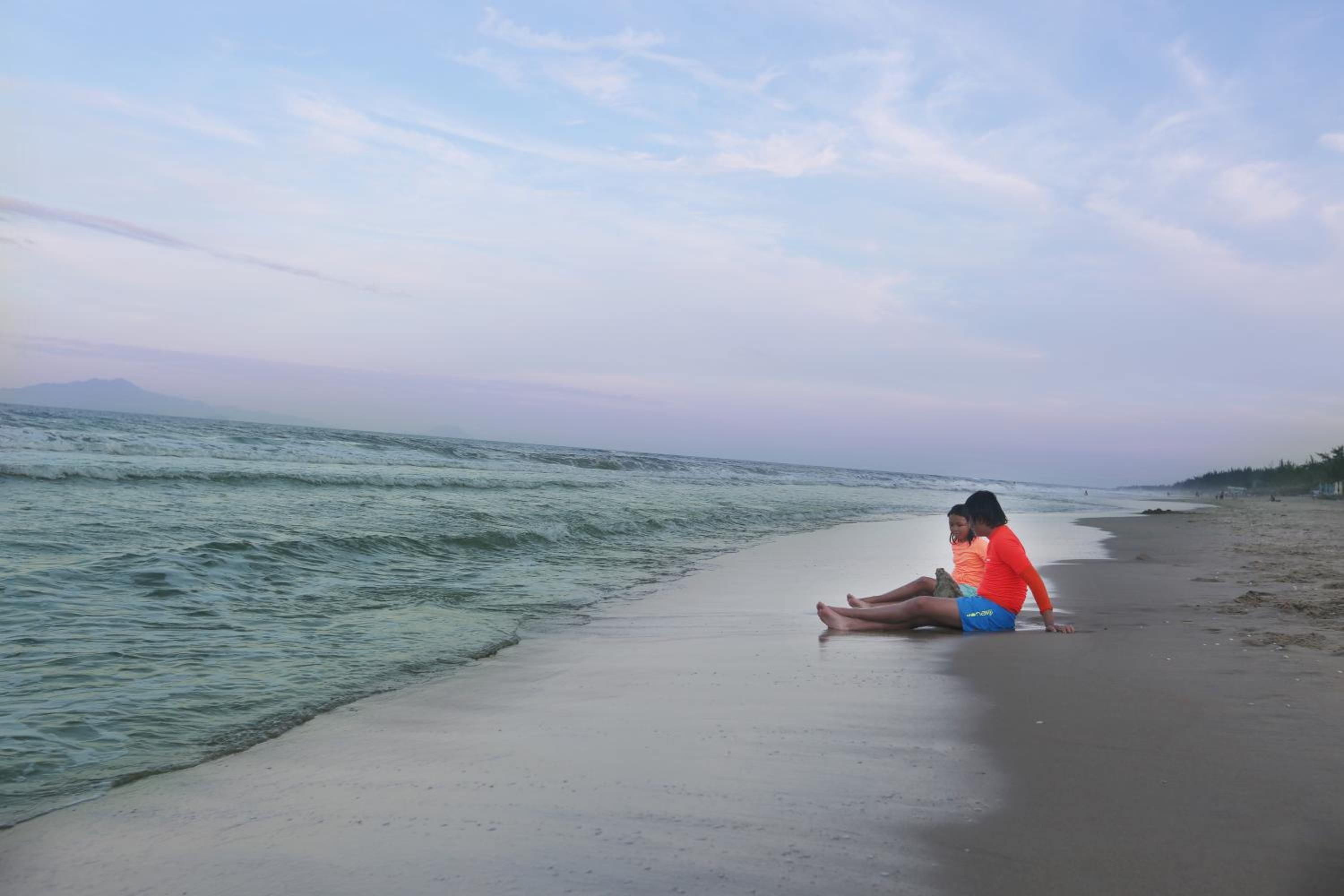 A beach with a person sitting on the sand near the water.
