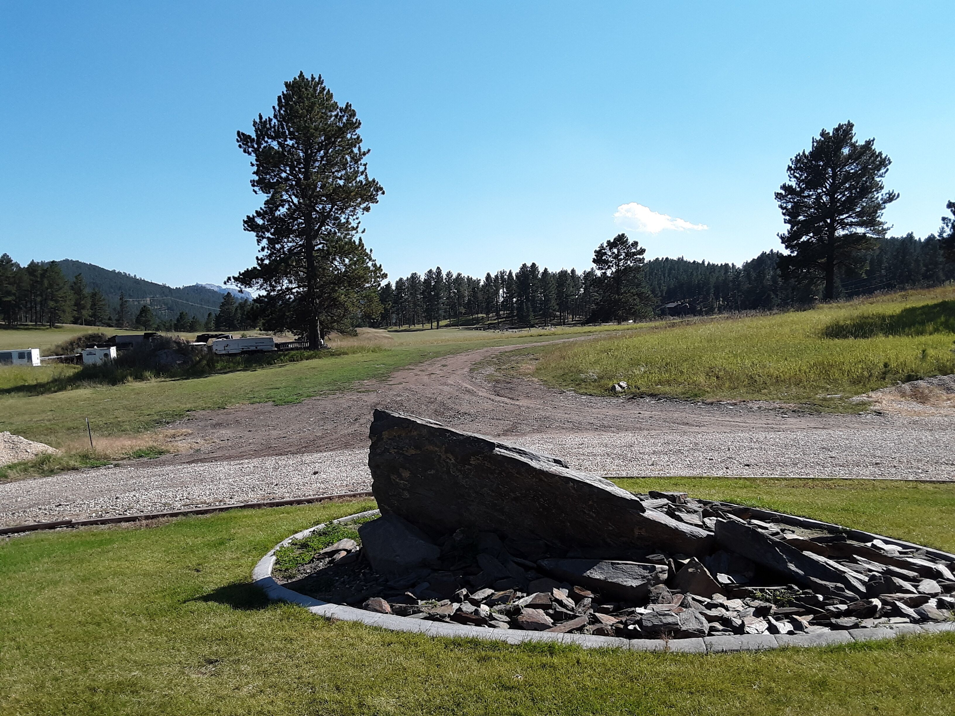 A rural landscape with a gravel path, a large tree, and a pile of rocks in the foreground.