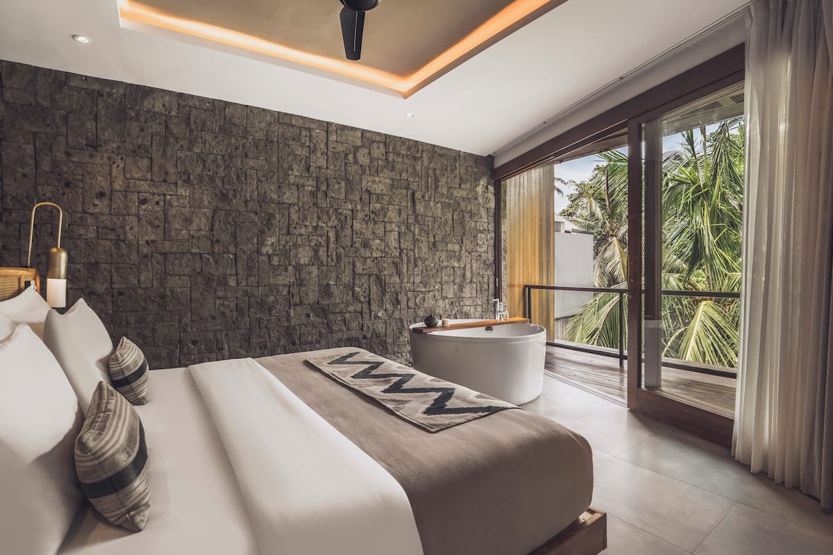 A modern bedroom with a large bed, a stone-textured wall, a round bathtub, and a balcony with palm trees.