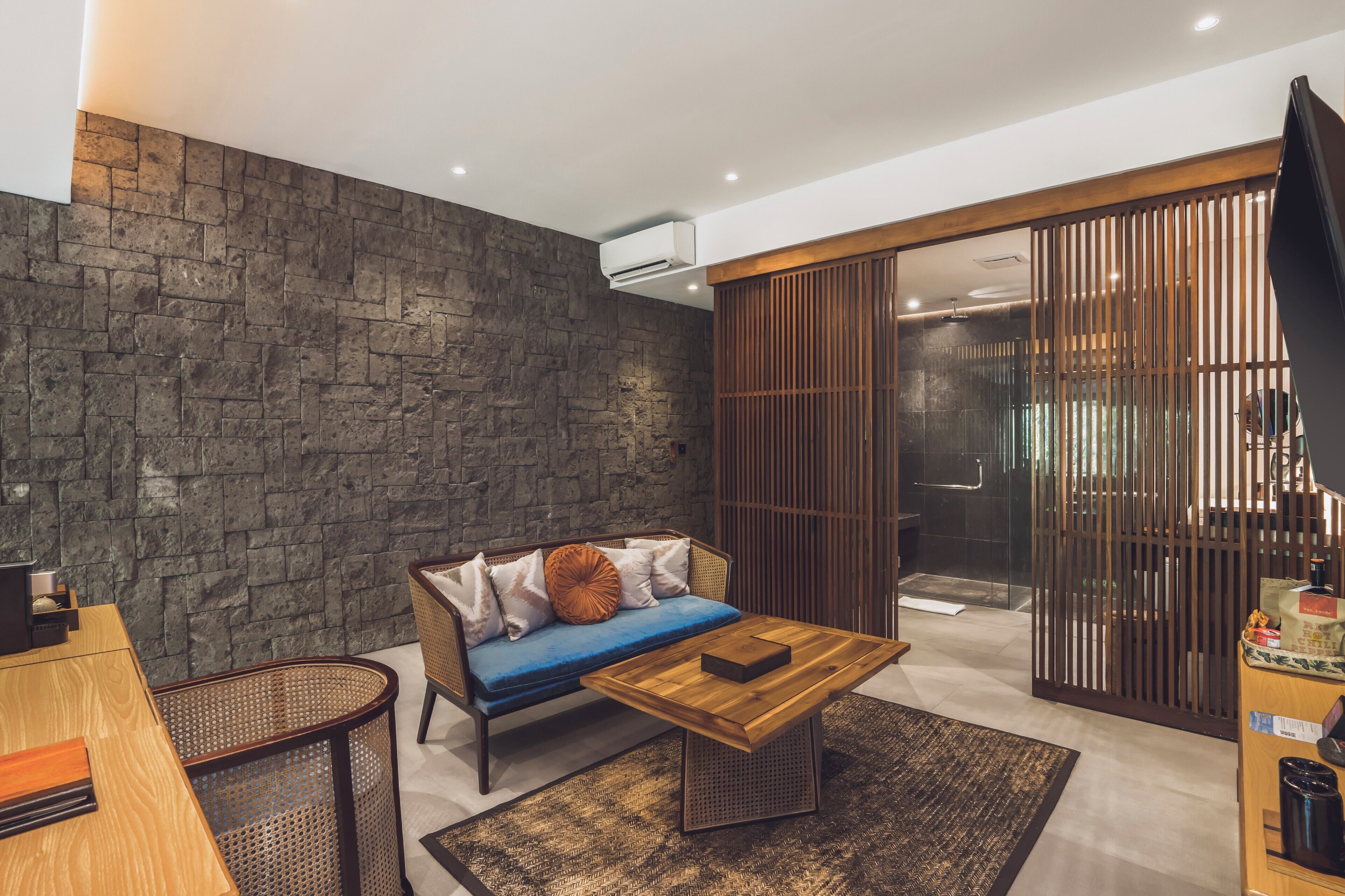 A modern living room with a stone wall, a sofa with cushions, a wooden coffee table, and a sliding glass door leading to a shower area.