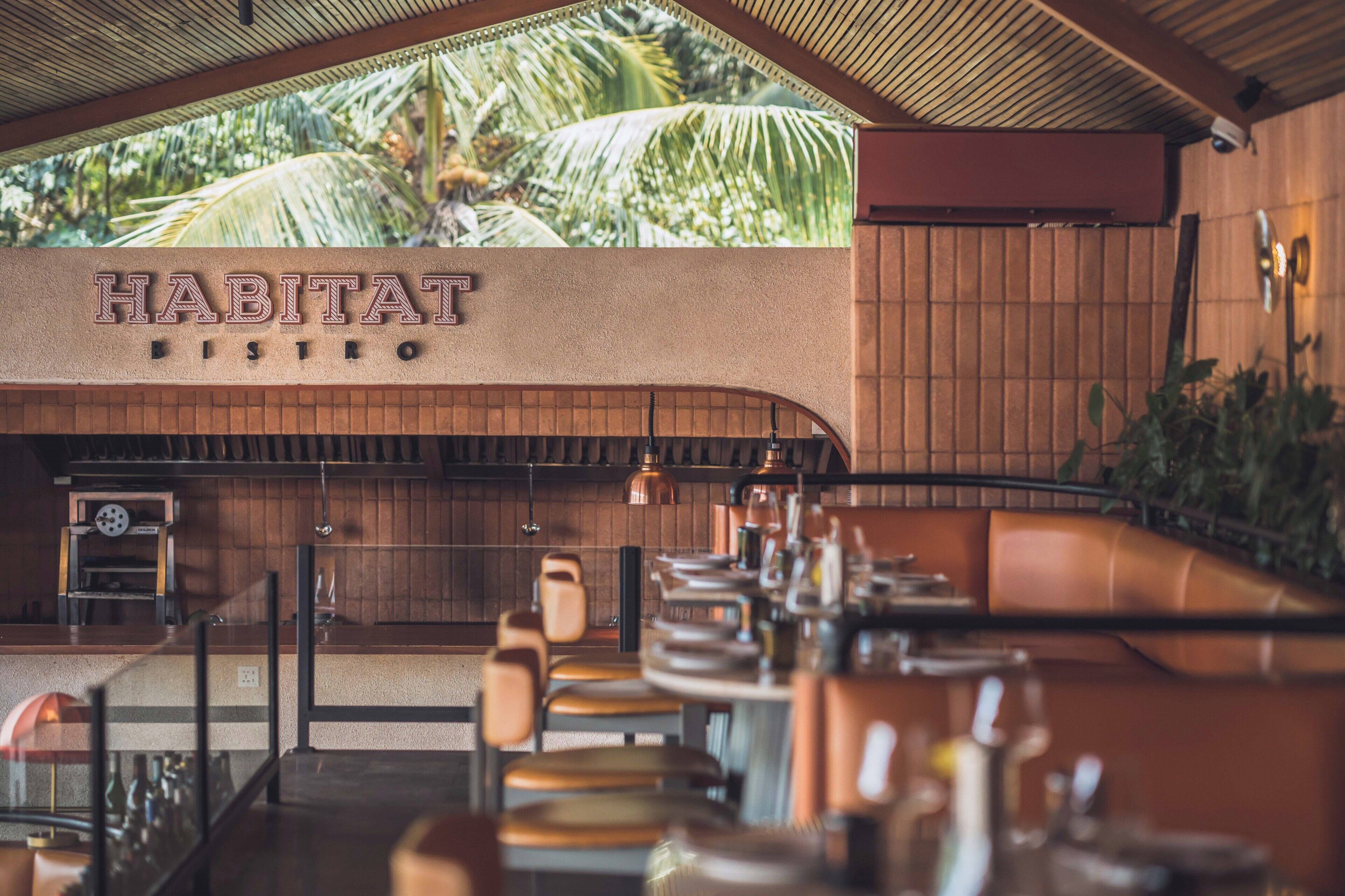 Ein Restaurantinnenraum mit einem Schild mit der Aufschrift 'HABITAT BISTRO', Holzvertäfelung und Blick auf Palmen.