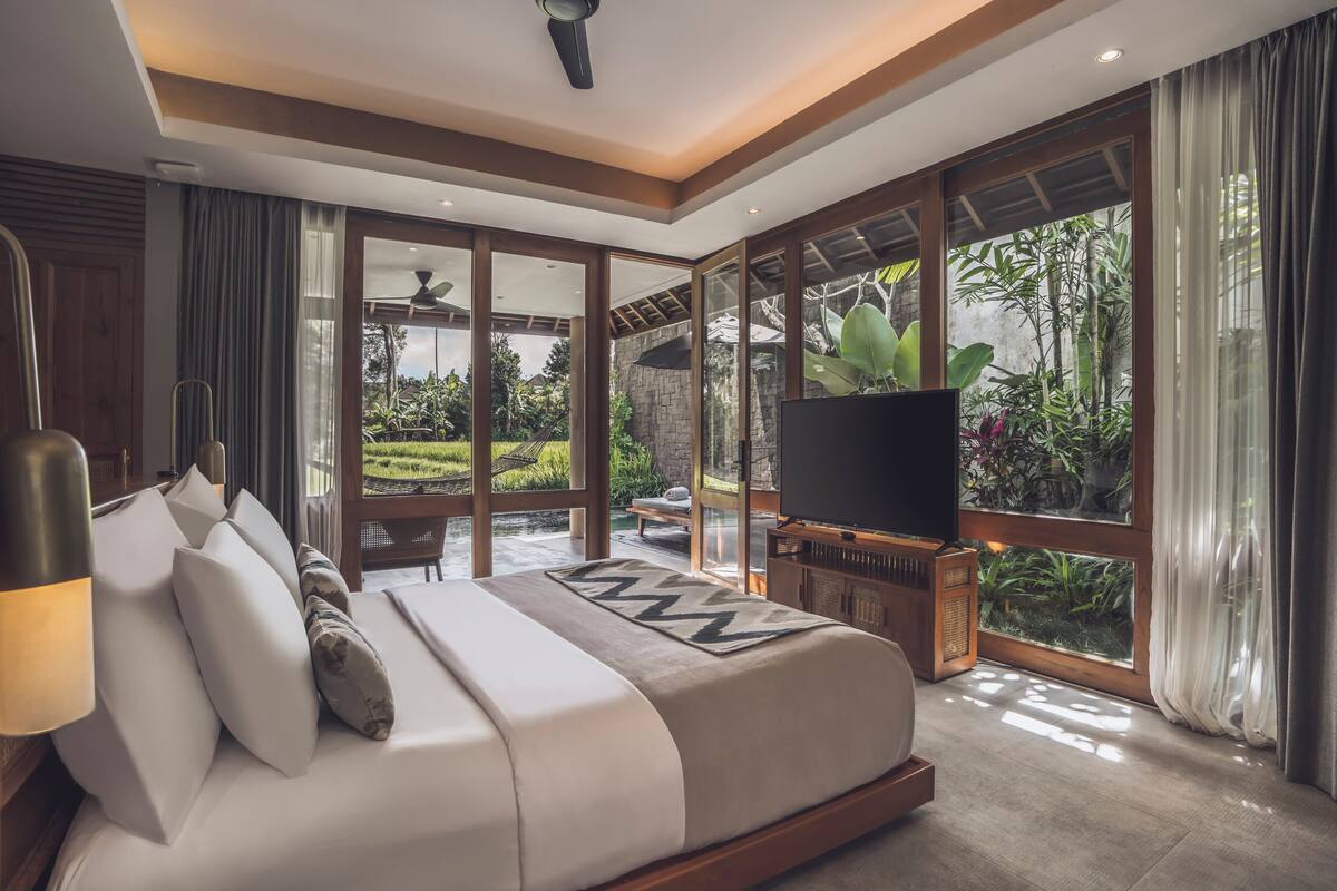 A spacious bedroom with a large bed, a television, and a view of a lush garden through large windows.