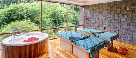 A spa room with a large window, a wooden bathtub, two massage tables, and a stone wall.