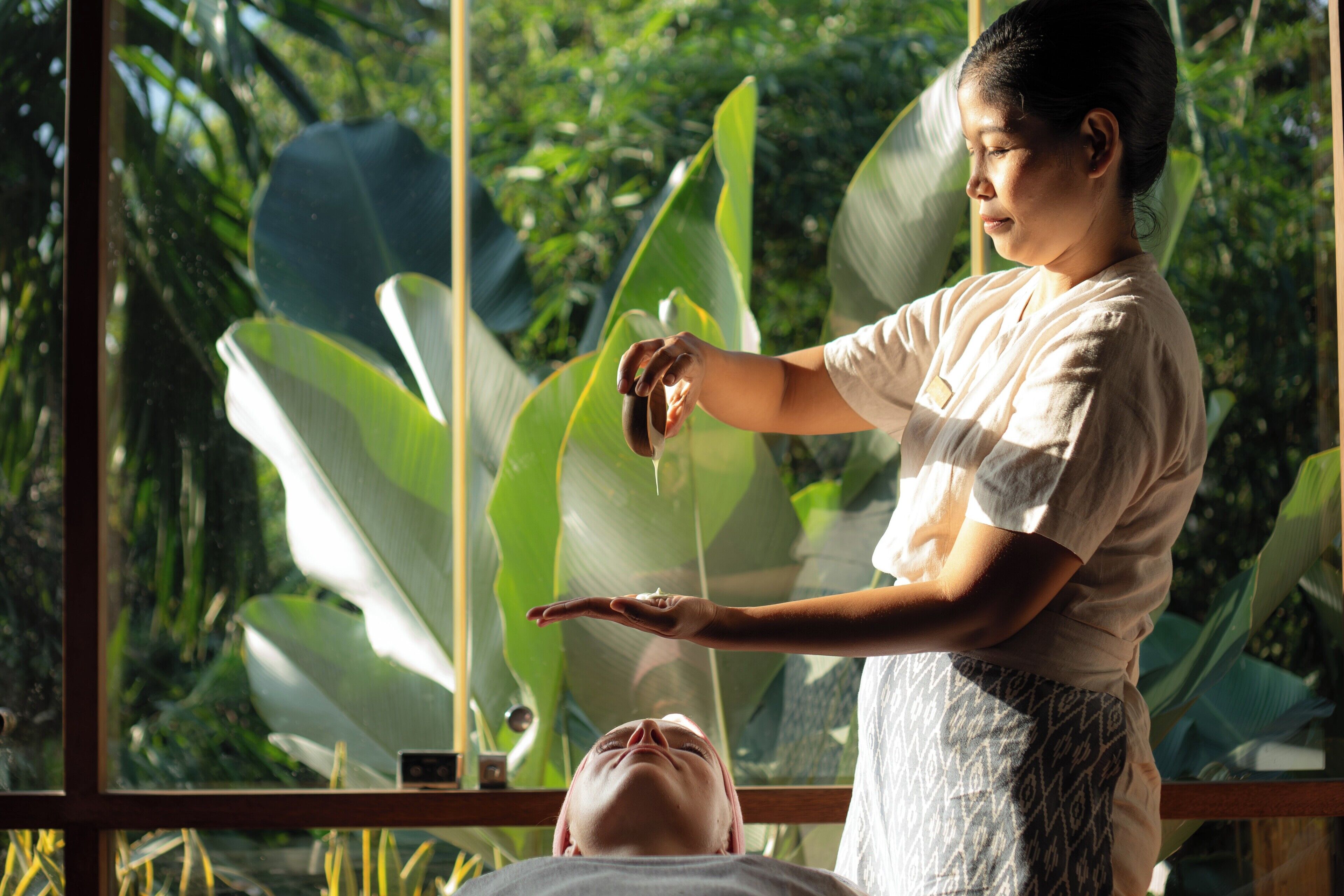 Une personne effectue un massage sur une autre, dans une pièce dotée de grandes fenêtres et agrémentée de plantes tropicales.