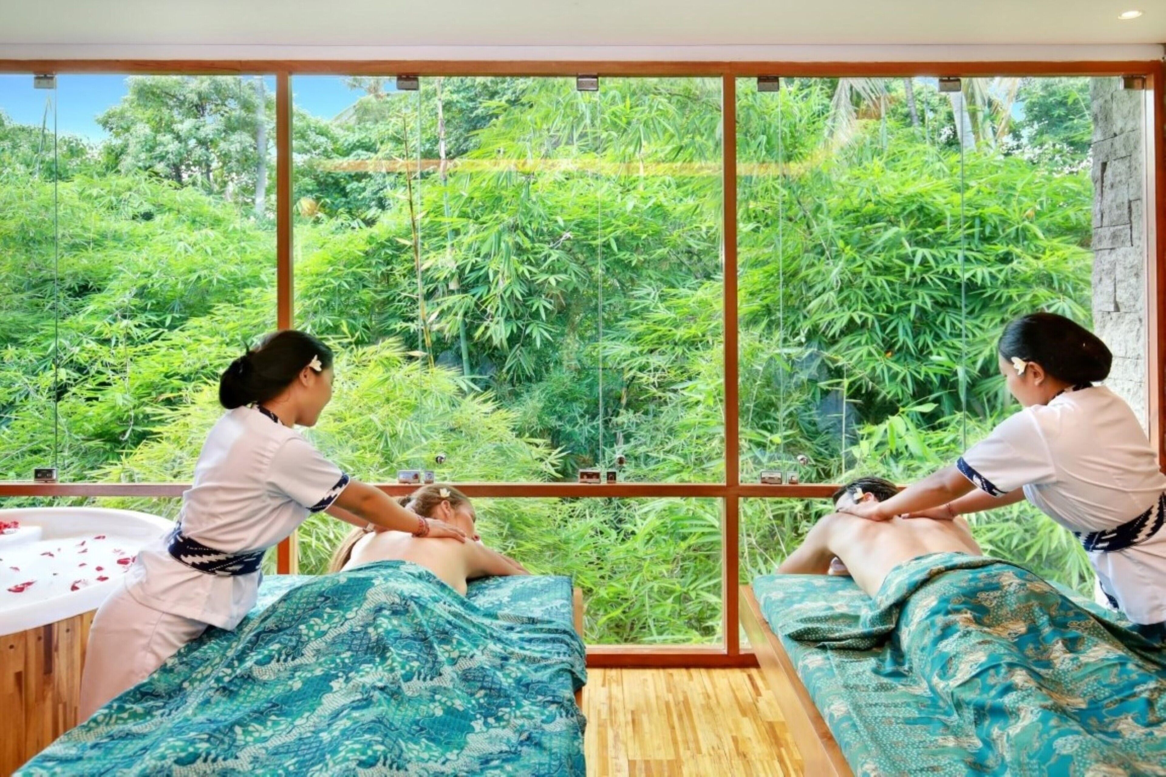 A spa room with large windows overlooking a lush green landscape, two massage tables, and a wooden floor.
