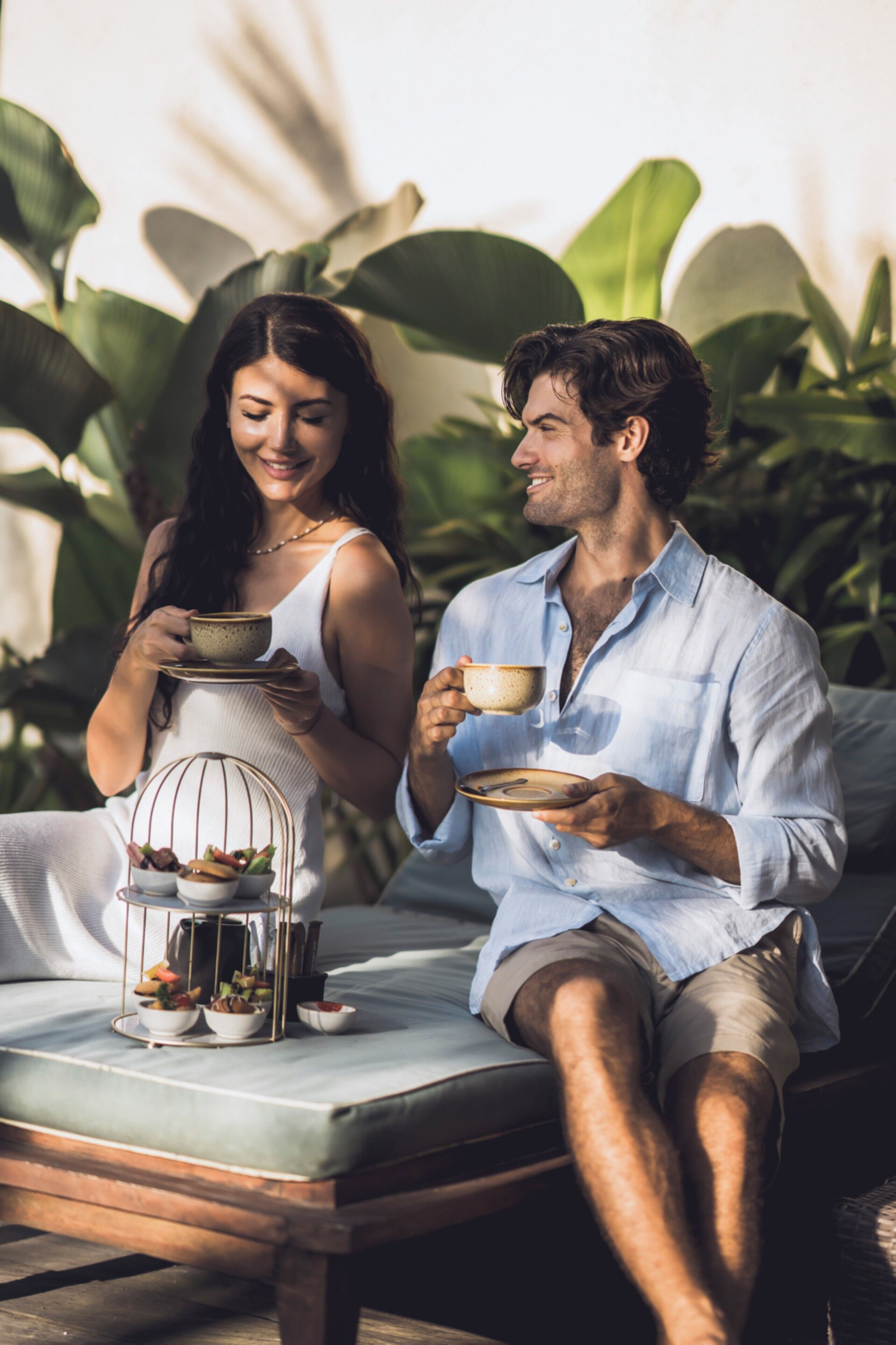 A couple sitting on a sofa with a tray of food and drinks, surrounded by greenery.