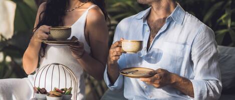 A couple sitting on a sofa with a tray of food and drinks, surrounded by greenery.