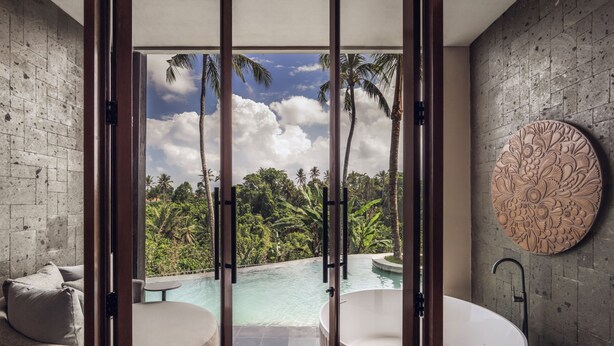 Un baño con bañera, un adorno redondo en la pared y vistas a una piscina y paisaje tropical.