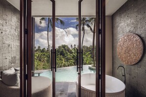 A bathroom with a bathtub, a round decorative wall piece, and a view of a pool and tropical landscape. - Kaamala Resort & Spa Ubud by Ini Vie Hospitality (Ubud)