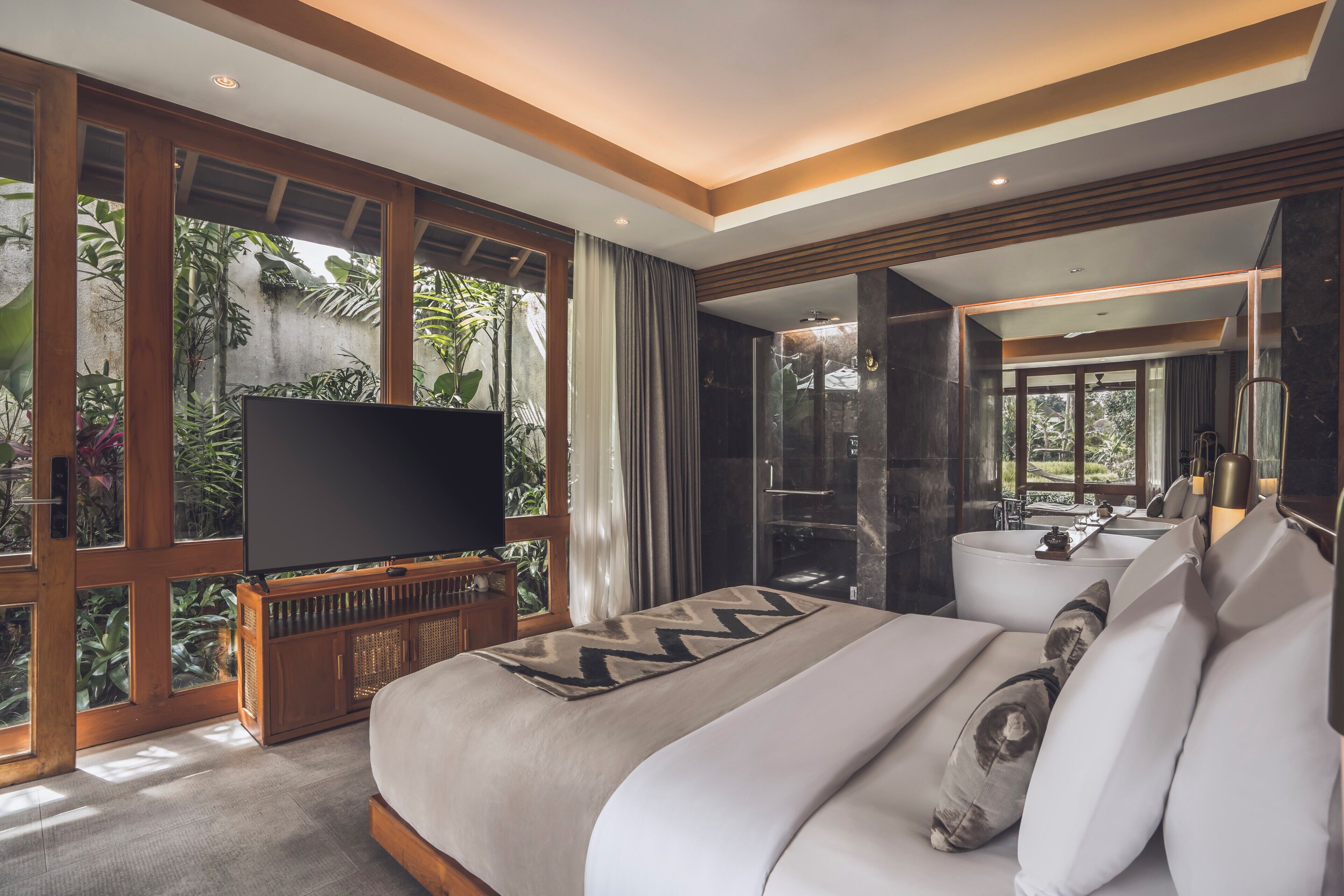 A modern hotel room with a large bed, a television, a bathroom with a bathtub, and a view of lush greenery.