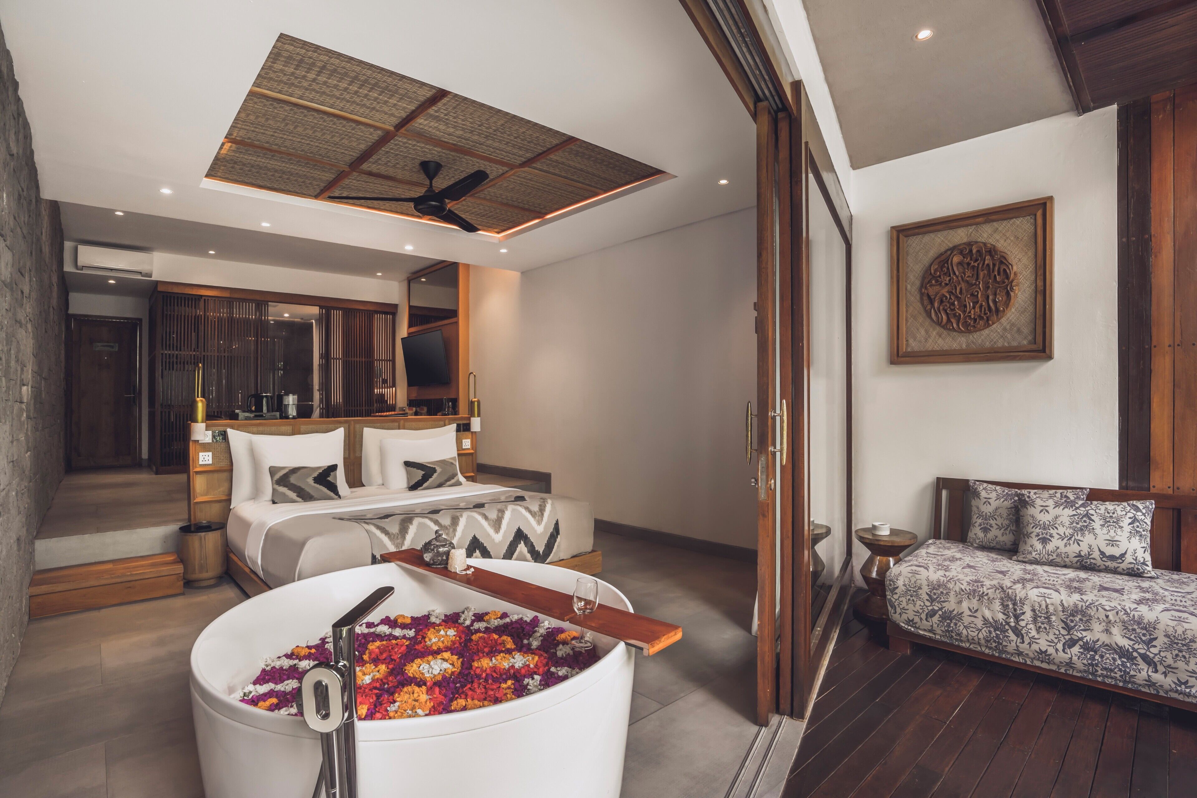 A modern bedroom with a large bed, a sitting area with a sofa, a bathtub with flowers, and a ceiling fan.